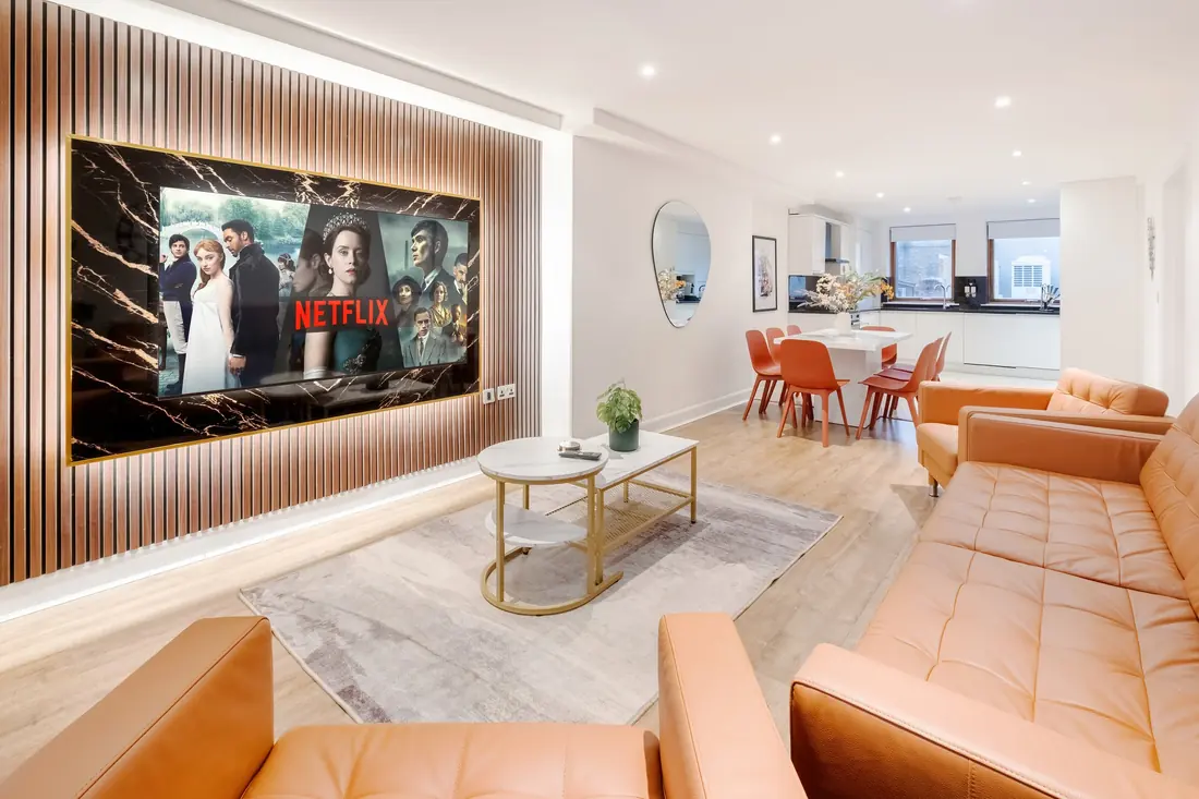 Living room with orange leather sofas, a 75 inch TV on a lit oak panel wall and the dining table beyond